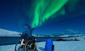 Usuaria de silla de ruedas y su hijo sentado en la nieve divisando auroras boreales en Tromso, noruega 